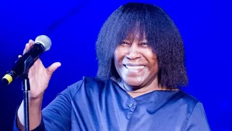 Getty Images A smiling woman with long dark hair and a blue shirt reaching for a black microphone on a stand