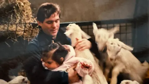 Sparks Family Maj Spark with his toddler daughter, Lucy. They are petting lambs in a farm setting.