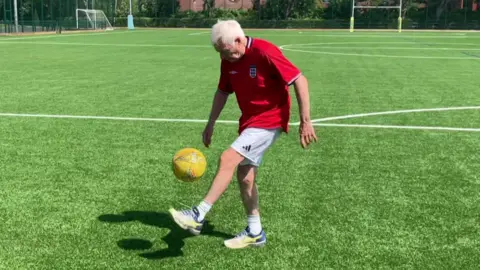 A person is standing on a green sports field, keeping a yellow football in the air with one foot. The field has goalposts, markings, and is surrounded by trees and red-brick buildings in the background. The person is wearing a red England shirt, white shorts, and trainers on a bright sunny day.