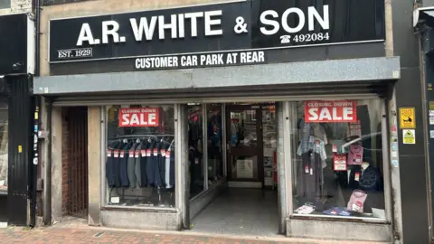 The entrance of AR White & Son. It has a black sign with the company name in white capitals above the door. Red signs in the window advertise a closing down sale, in front of a variety of men's suits and shirts.