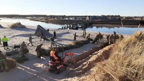 Moray Council Orange mini digger working on a trench on a beach, while RAF personnel in camouflage help move trees.