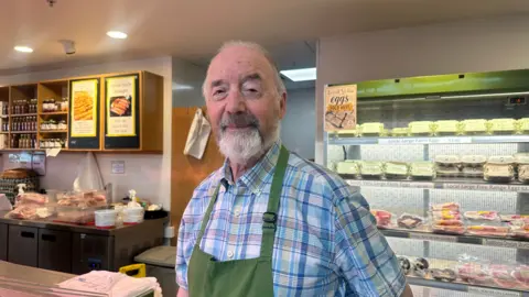 A man with a grey beard. He is wearing a checkered blue shirt and green apron. He is standing inside a deli with shelves of packaged meat behind him.