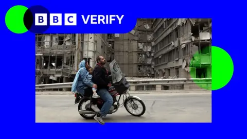Man, woman and child on a motorbike passing in front of destroyed facade of the Gandhi hospital in Tehran