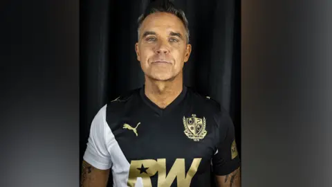 Robbie Williams, a man with grew spiked-up hair, is looking directly at the camera. He is standing against a black background, wearing a black white football shirt with the club logo and the letters RW in the centre in gold lettering.

