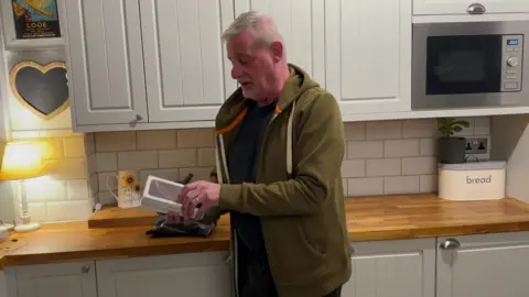 Tony Deeley in a green jacket holding an iPhone box. He is standing in his kitchen.