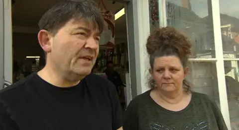 A man and a woman stand outside the open door to a shop. The man has dark hair and wears a black T-shirt. The woman has dark hair in a high ponytail and is wearing a grey T-shirt with a diamante butterfly design.