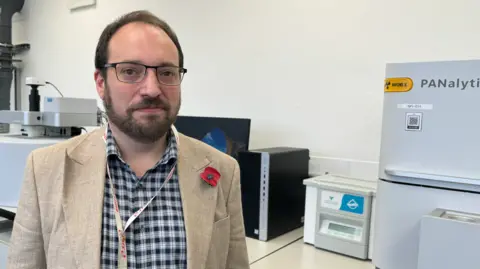 Simon Middleborough portrait, wearing a checked black and white shirt with a light coloured jacket, wearing a remembrance poppy - situated in a laboratory with scientific machinery behind him