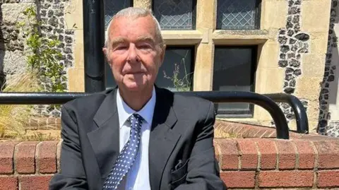 Keith Gould is wearing a suit and sitting outdoors beside a stone building with large windows and brickwork in the background.
