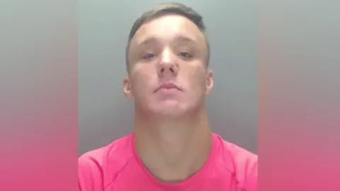 Mugshot of Brandon Welch, he has short fair hair and is wearing a pink t shirt.