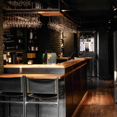 Emilia Boscarelli The bar area at the restaurant with dark walls and dark brown wooden flooring. Two black leather bar stools are tucked under the bar at the front and wine glasses hang from an overhead rack all around.