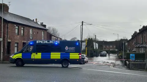 Handout A large dark blue Lancashire Police van is parked at the entrance to a street lined with houses, which is partially submerged