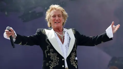 Rod Stewart is standing on stage wearing a black jacket with stitched flowers. He is spreading his arms towards the crowd and has a microphone in hand. His shirt has a low neckline and he is wearing about three necklaces.