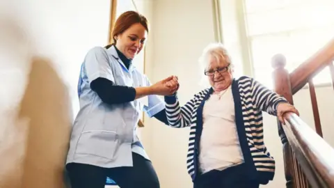 A care worker helping an elderly woman walk down a staircase. The carer, on the left, is wearing a light-blue jacket and dark trousers. She is smiling and holding the older woman's hand. The older woman has white hair, is wearing spectacles and has a look of concentration on her face as she takes the steps. She is holding the banister with her left hand.