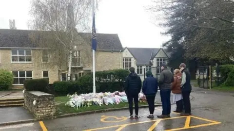 A group of five people stood next to each other, in front of flowers that were been laid by a flag, at half mast, outside the front of a stone-built school building