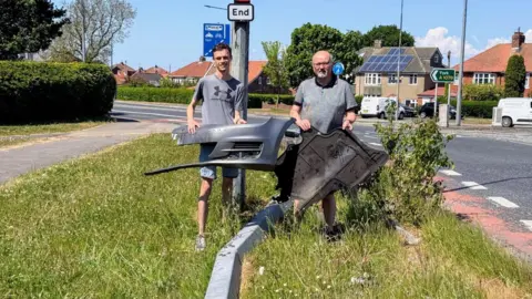 York Liberal Democrats Two men hold up debris from a car, next to a bent crash barrier.