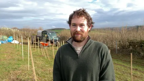 The picture shows a person standing in a field where rows of wooden stakes stretch out behind them. They wear an outdoor fleece and stand in front of a muddy track where a small utility vehicle and a trailer are parked. Bags and tools sit off to one side, with low winter sunlight breaking through the clouds and rolling hills in the distance. 