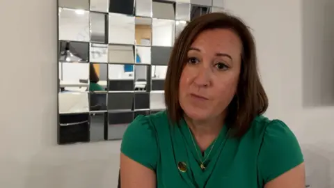 Alex has shoulder length dark brown hair. She is wearing a green top.