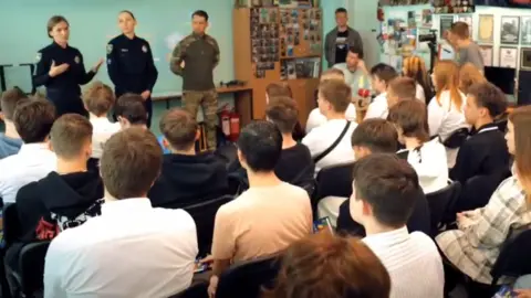 SBU A uniformed female SBU officer stands in front of a class of teenagers in a school in Kyiv, delivering a talk warning about the dangers of being recruited online to carry out sabotage against their own country. Beside her are two other SBU officers, one male and female, along with a teacher. The talk is being filmed by a camera operator from the side of the class.