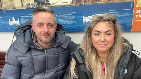Michael and Lucy Gittins pose for a picture while sitting on a passenger bench. A board behind them details the area's history.