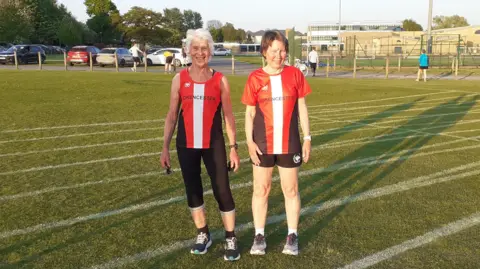 Ruth Fulford Ruth Fulford (left) and Pam Wheeler in red and white Cirencester athletics tops and black shorts and leggings. They are smiling at the camera, standing on a grass athletics track. There is a car park in the background.