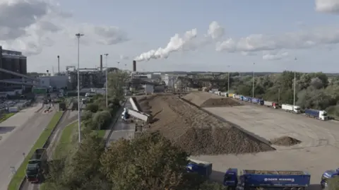 A factory with smoke billowing from a chimney with a queue of lorries in front of it depositing sugar beet