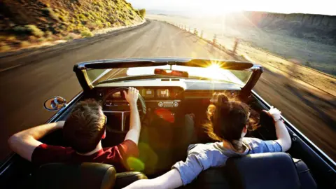 A young man and woman in a convertible car driving along a mountain road towards the sunset. She is driving and has her arm around the back of his seat.