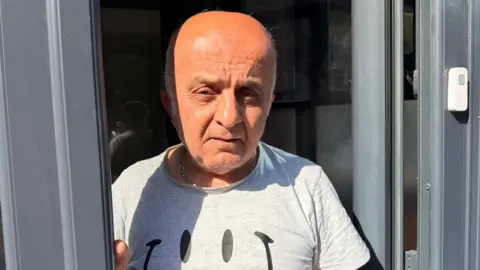 A man with a grey T-shirt with a smiley face on it. He is mostly bald with grey hair on the sides of his head. he is looking at the camera with a stern face.