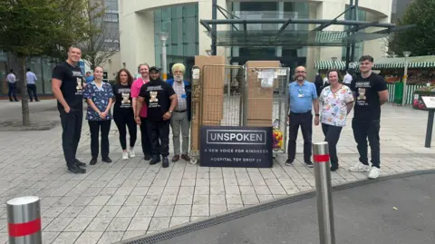 Atiq Rehman Atiq and his team of volunteers and some hospital staff members pose outside a hospital with boxes of toys. At the front of the boxes a sign is propped up which reads "Unspoken: A New Voice For Kindness Hospital Toy Drop 24". 