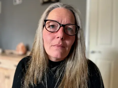 Kellie Shone has long greying hair, she wears black-framed glasses and a black top. 