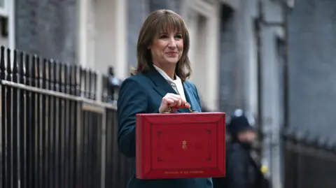 Una mujer con el pelo hasta los hombros se encuentra frente al número 10 de Downing Street, junto a una barandilla negra. Vestía un traje azul marino y llevaba un maletín rojo mientras sonreía.