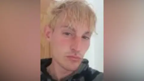 Andrew Haire, a man with short, blonde hair, looks to the camera in a close-up photo.  He is wearing a grey and black hooded and has a bit of light stubble on his upper lip.