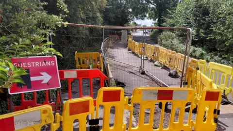 The Castle Cary sinkhole on the B3152 Station Road. The area around the sink hole is closed off with metal fencing and yellow and red bollards. 