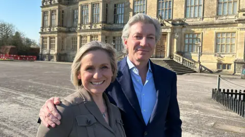 Lord Bath stands before Longleat House beside the BBC's Amanda Parr. Both wear dark jackets and are smiling at the camera on a bright sunny day with blue skies behind them.