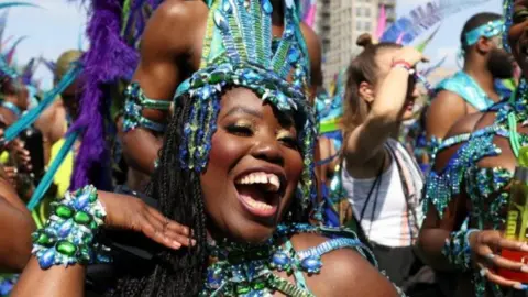 PA Media A woman wearing a blue and green sequinned head dress as well as a beaded costume smiles while other people stand in the background at Carnival