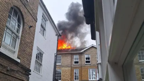 Flames and heavy smoke coming from the roof of a brick building in a built-up area.
