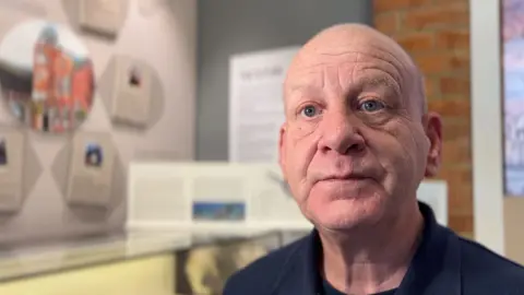 Dr Paul Davis is in the right hand of the frame in focus. In the background out of focus is the display of the museum's exhibit, but not much detail can be made out. 