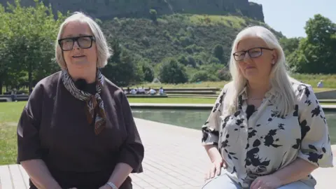 Two women sit by a plaza with a water feature and hill in the background. Cynthia Guthrie wears a brown top and dark glasses, and Allison Graham wears a patterned white and black shirt with dark glasses. Both have fair hair 