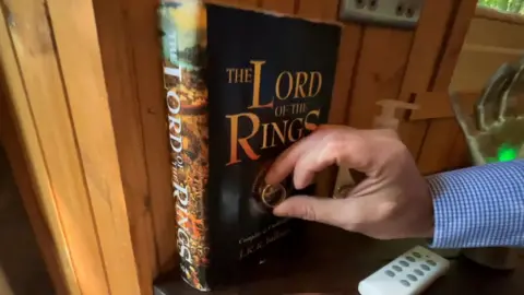 A black hardback copy of the lord of the rings against a wooden wall. A hand is placing a ring against the front of the book.