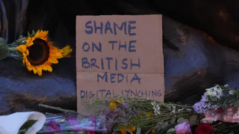 PA Media A cardboard sign with the words "shame on the British media. Digital lynching" among floral tributes.