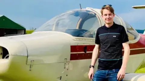 A man in a black polo shirt with white stripes stands in front of a light aircraft, smiling. It is a sunny day.