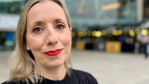 JO THEWLIS/BBC Sarah Taylor has shoulder-length blonde hair and is wearing a black top and cardigan. She is smiling into the camera and photographed outside a glass-panelled building. 