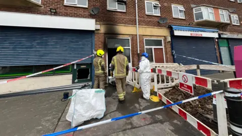 A police cordon set up outside the front of a terraced building with flats and shops. Firefighters and officers dressed in white forensic outfits are looking at the entrance to a flat, which has been scorched by a fire.