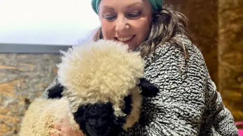 A woman wearing a green hat with a sheep. There is a brick wall behind them. 