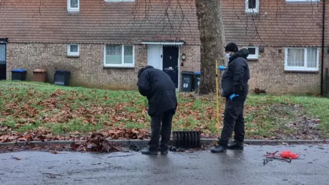 Dawid Wojtowicz/ BBC Two police officers dressed in black with masks and blue rubber gloves look into an opened drain by a kerb in a housing estate.