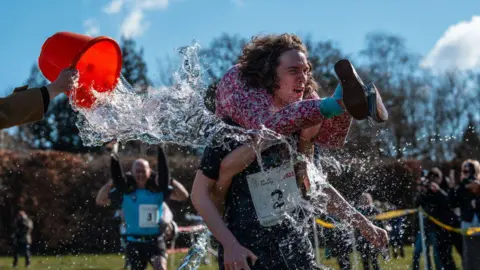 Getty Images A man carrying a partner on their back with their legs wrapped around their shoulders. A person is throwing a bucket of water over them.