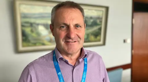 A man wearing a white and red chequed shirt and a blue lanyard, smiling to the camera inside an office with a landscaped painting of the Shropshire town of Ludlow behind him 