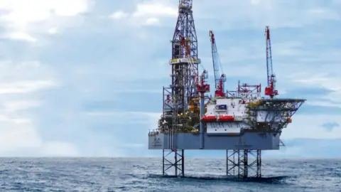 A rig sits in the sea protruding above the horizon with a blue sky with clouds. Three red cranes and a tall skeletal metal structure rise high from a sturdy grey deck.