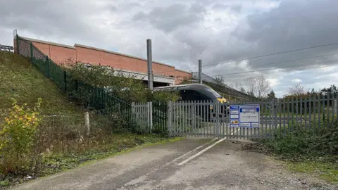 A bridge going over a railway with a train going underneath it. Next to the bridge, a road can be seen crossing the track so it's evident there used to be a level crossing there, but now it's fenced off.