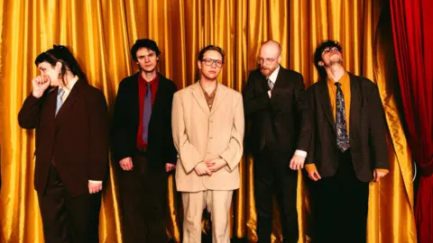 A promotional photo of The Howl & The Hum. The five band members made up of one woman and four men are standing in front of a large golden curtain. They are all wearing dark suits with ties, except for the man in the middle who is wearing a beige suit with no tie and his top shirt button is undone, he is also wearing retro spectacles on his face.