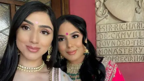 Jussi Sidhu Two British Indian women taking a selfie. Jussi on the left is wearing a light pink bindi and gold and silver big Indian earrings and a necklace. Her sister Kiran is to the right of her wearing a dark pink bindi on her forehead. She has has gold silver and green big indian style earrings and a necklace. She is a wearing a bright pink sari.